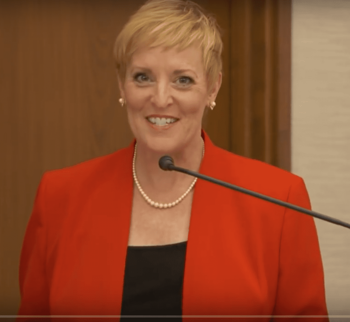 A woman in red jacket standing at podium with microphone.